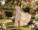 A woman wearing a cream floral midi dress with flutter sleeves and a V-neckline, walking in a sunlit garden with blooming trees and white and pink flowers.