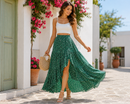 A woman walking outdoors in a white crop top and a flowing green patterned maxi skirt, holding a woven straw bag. The setting features white stucco buildings, a light green door, and bright pink bougainvillea flowers.