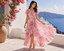 A woman wearing a pink floral high-low midi dress with flutter sleeves, standing on a stone patio with a scenic blue sea and white buildings in the background.
